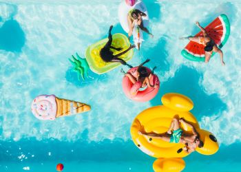 Attain on 5th apartment homes with Five people relax on colorful pool floats shaped like food in a bright blue swimming pool, viewed from above.
