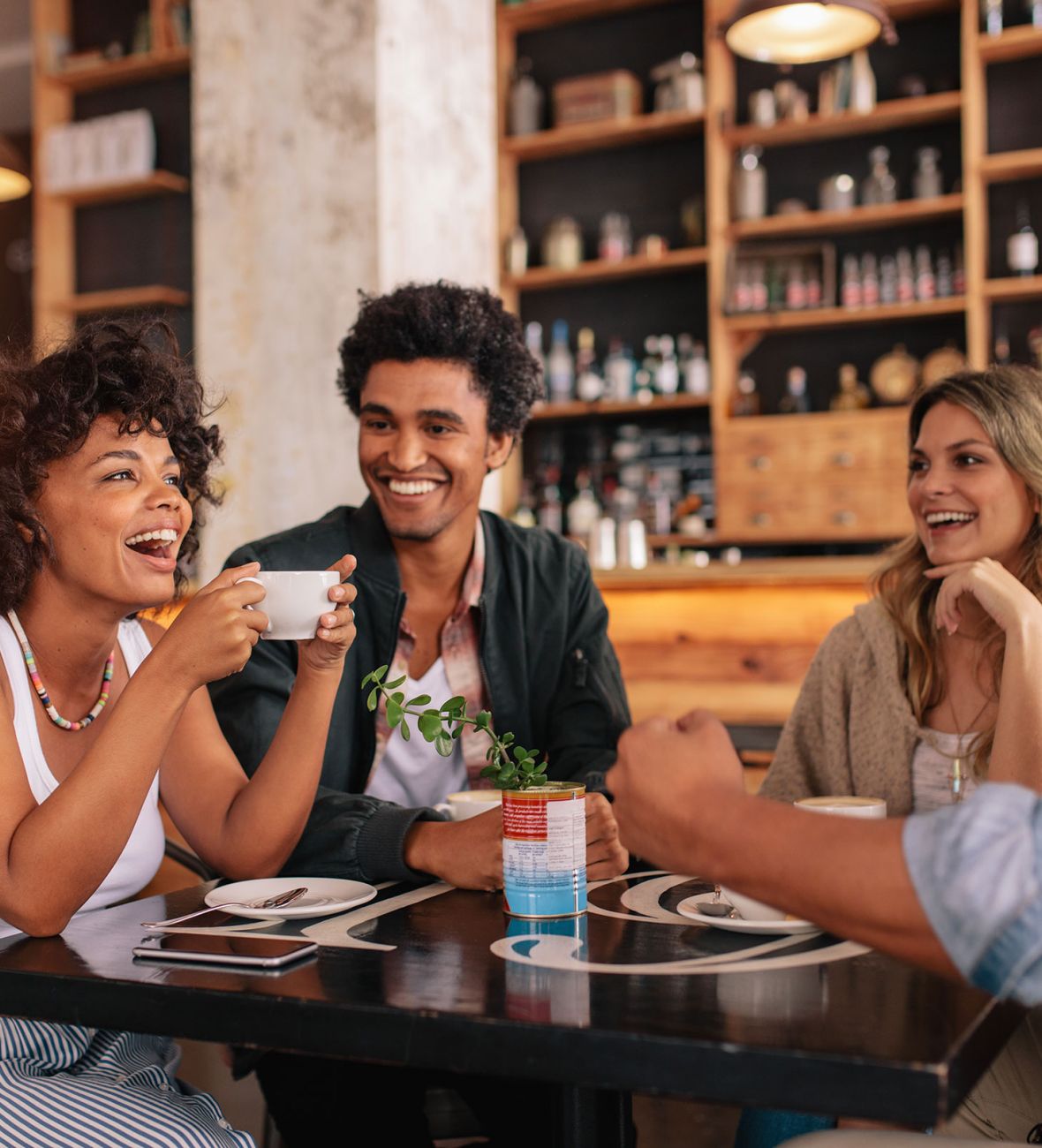 Attain on 5th apartment homes with Four friends sit at a café table, laughing and enjoying drinks together in a cozy, warmly lit setting.